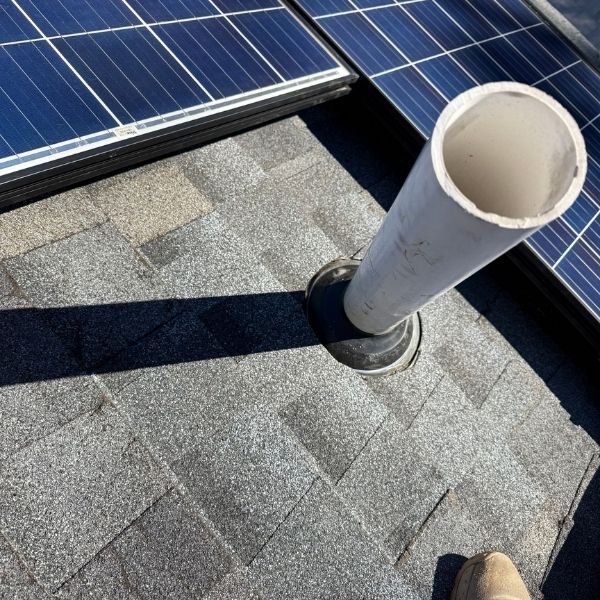 A white PVC plumbing vent pipe extending from a grey shingled roof next to a solar panel.