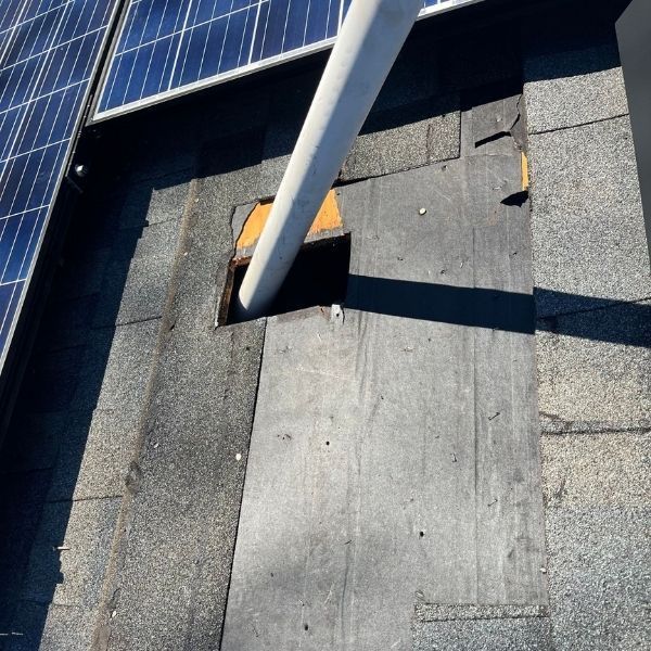 A white roof vent pipe protrudes through a square hole in damaged asphalt shingles on a roof near solar panels.