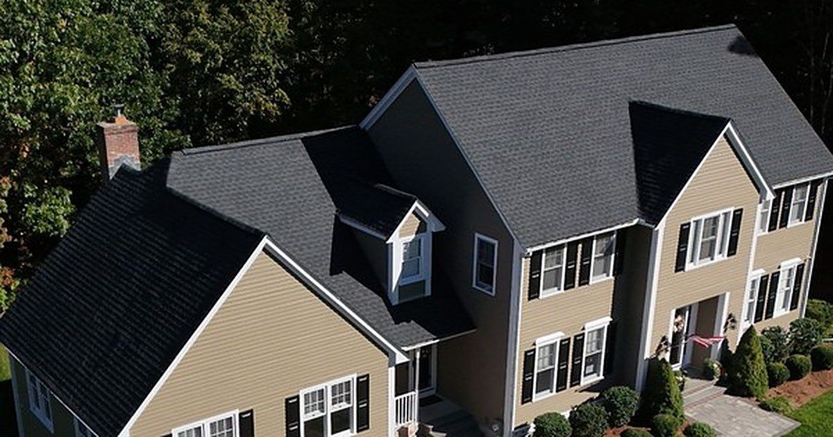 Aerial view of a beige suburban house with a dark roof and dormer windows, surrounded by trees and shrubs