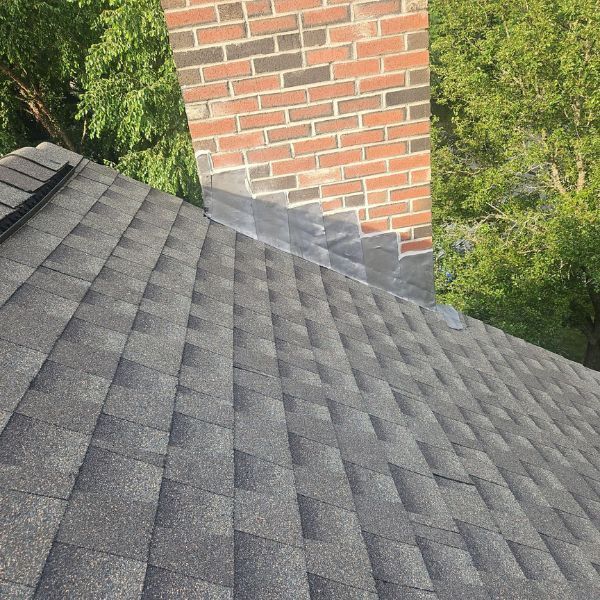 Roof with gray shingles, brick chimney, and flashing. Green trees in the background.