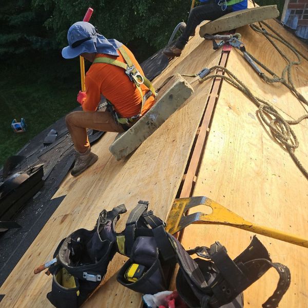 Roofer on a roof, wearing safety gear. Hammering with a red hammer, securing plywood. Green grass, blue sky.