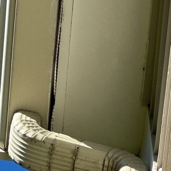 White downspout connected to a building with light yellow siding; crack in the wall.