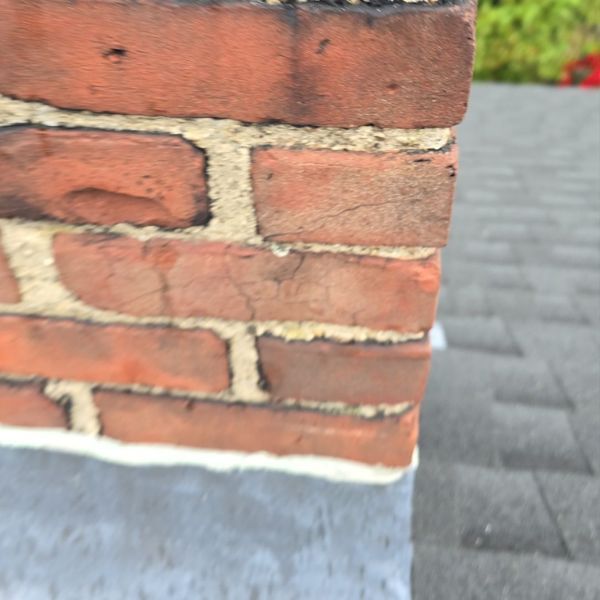 Close-up of a brick chimney on a gray roof, showing cracked mortar and a metal flashing.