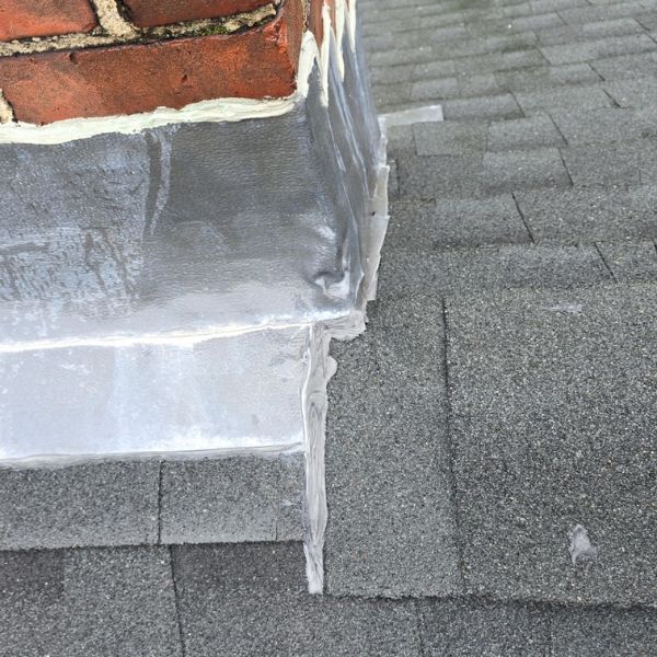 Close-up of chimney flashing on a shingle roof; gray shingles, brick chimney, and metal flashing.