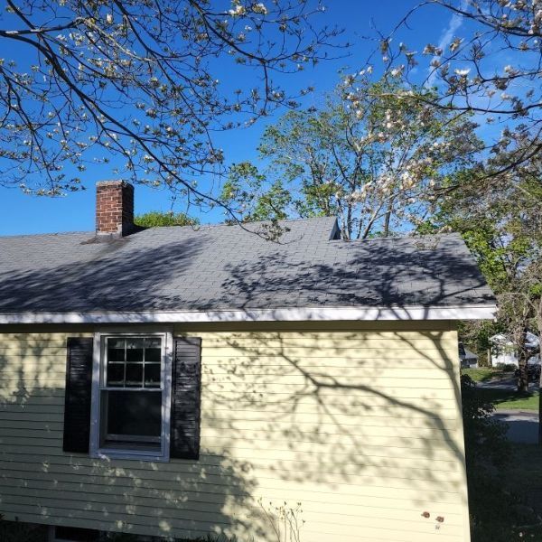 Yellow house with a gray roof and brick chimney under a blue sky, tree branches casting shadows.