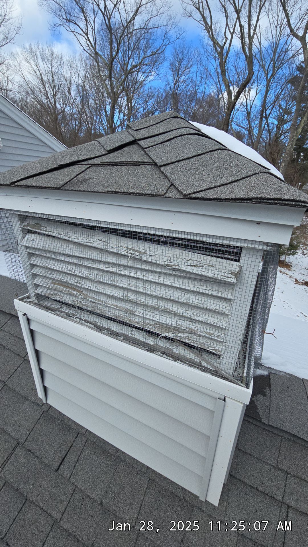 Damaged roof vent on a house, with broken gray shingles and white siding. Winter setting, some snow visible.