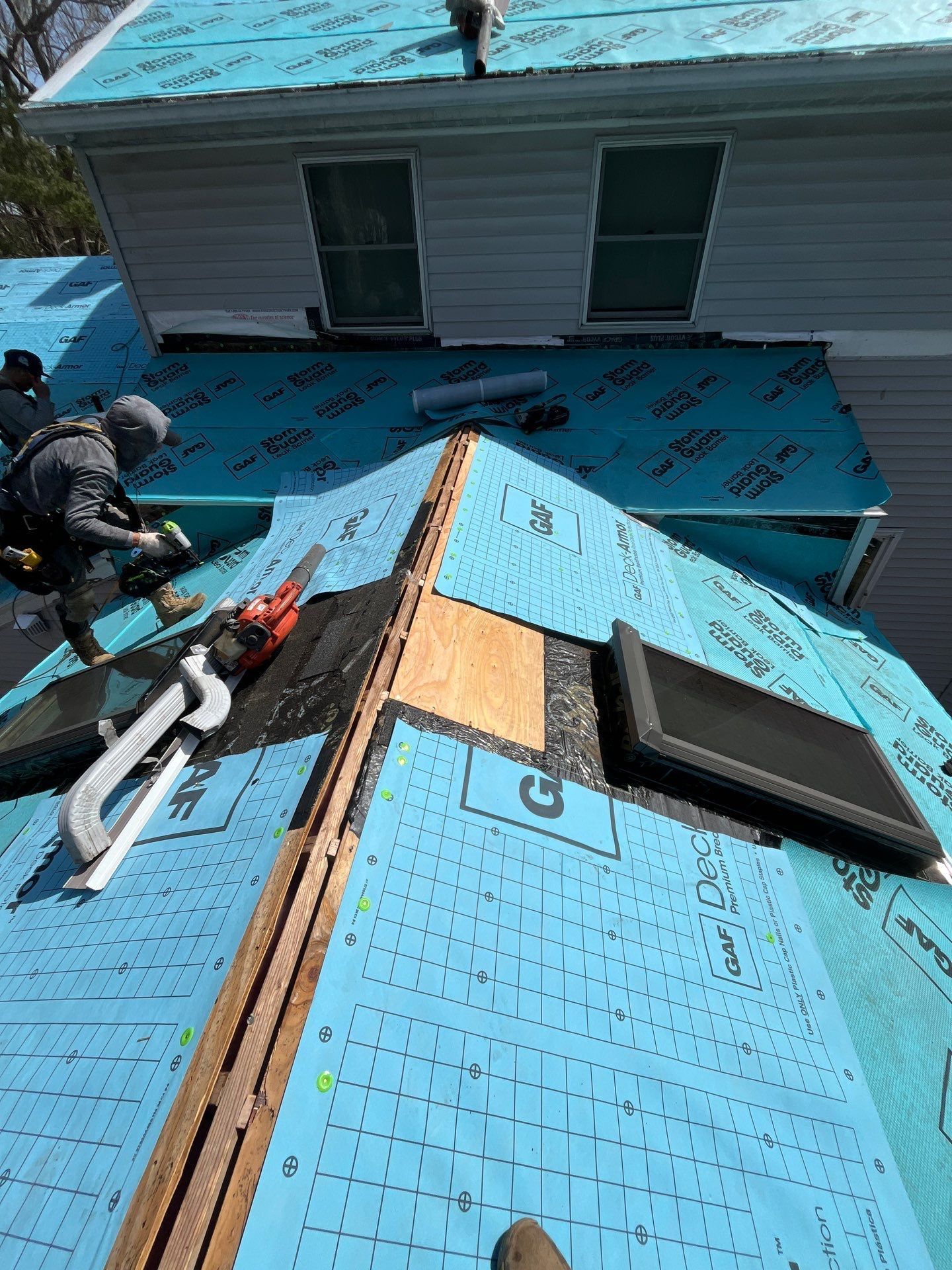 Roofers working on a house with blue roofing underlayment, one using a saw.