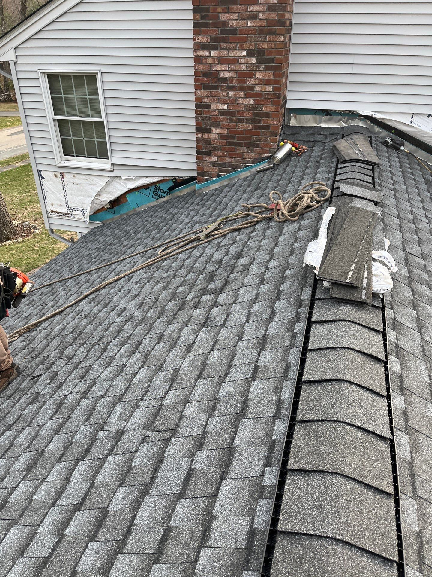 Rooftop with gray shingles; a rope and damaged sections are visible near a brick chimney and white siding.