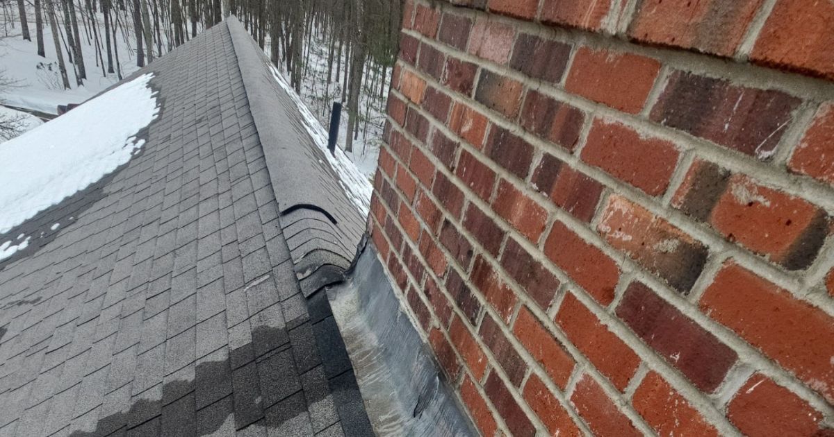A view from a roof showing a brick chimney with metal flashing meeting the gray shingled roof and patches of snow.