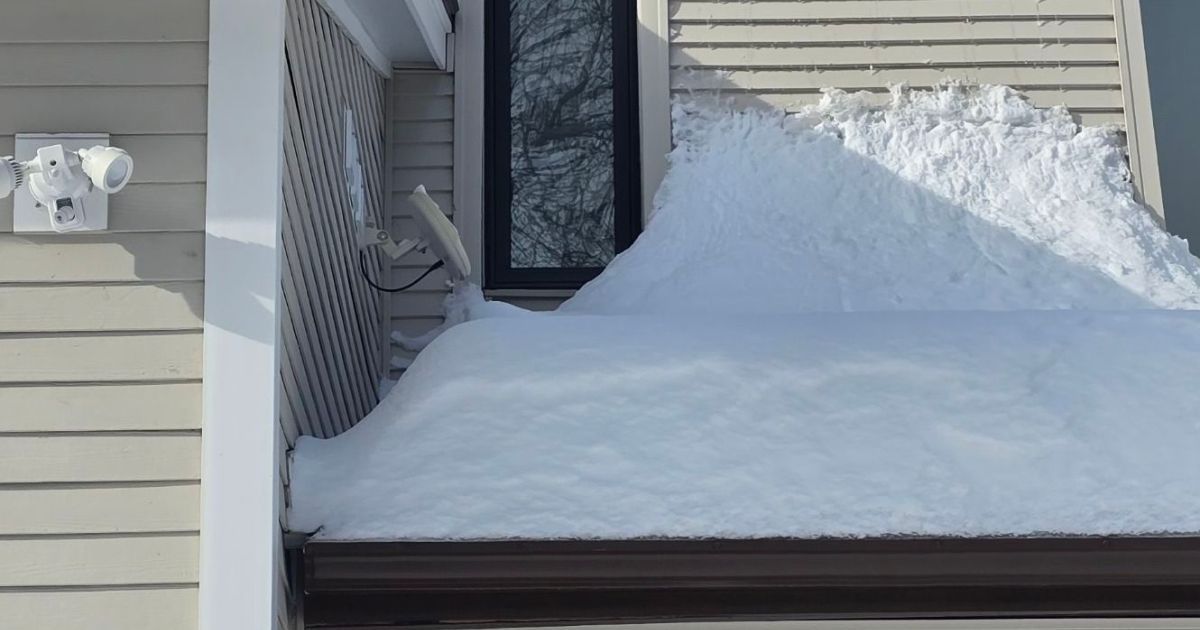 A thick layer of snow covers a roof below a window, with a satellite dish partially buried on the left side.