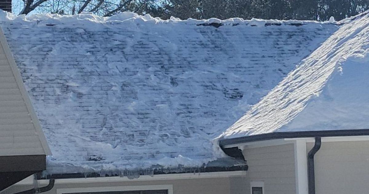 A snow-covered house roof with visible shingles, icicles along the gutters, and bright sunlight hitting the snow.