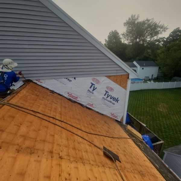 A worker in blue coveralls is installing siding on a house roof covered in Tyvek, green grass, and white fence.