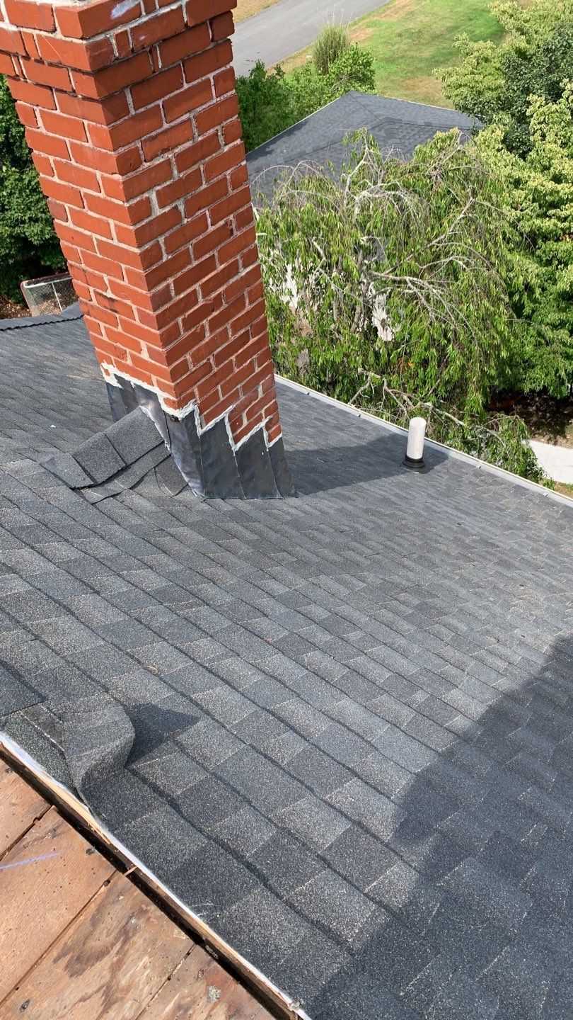 Brick chimney on a dark shingled roof with a small vent pipe, surrounded by trees.