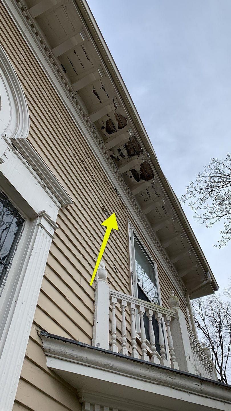 Exterior of a building with peeling paint, ornate trim, and visible roof damage. Yellow arrow points to the wall.
