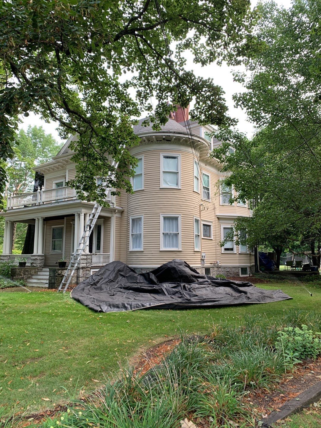 Victorian house with a wrap-around porch and a black tarp on the lawn for a renovation project.