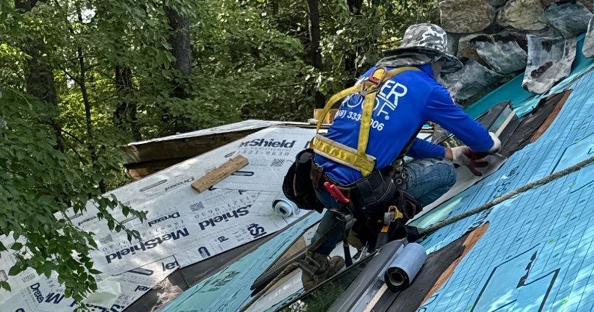 Roofer in blue shirt and safety harness installing shingles on a roof.