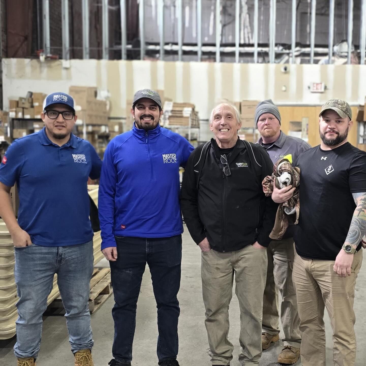 Five people stand together in a warehouse, some wearing hats and branded apparel, with a small dog held in the center.