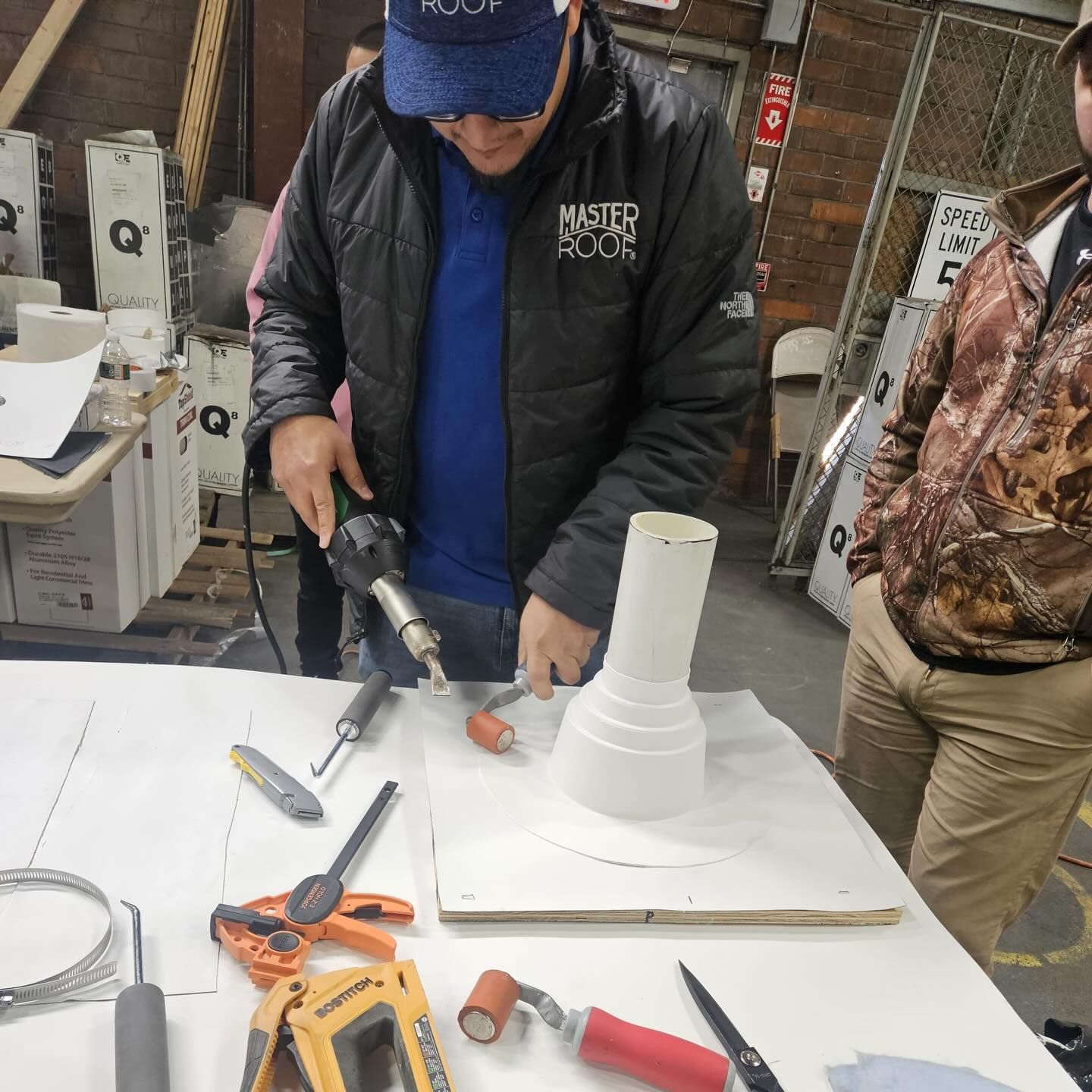 A person in a black jacket uses a heat gun and roller on a white pipe flashing assembly on a workbench in a workshop.