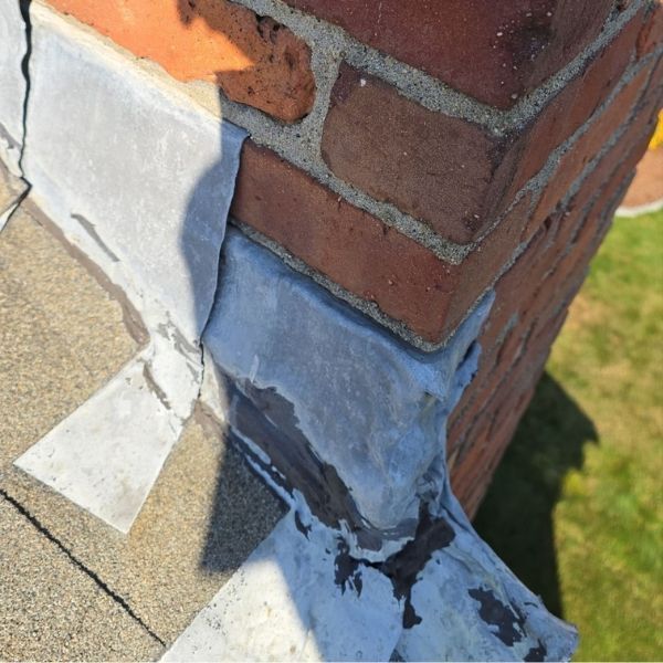 Chimney flashing with peeling sealant on a brick chimney and asphalt shingle roof.