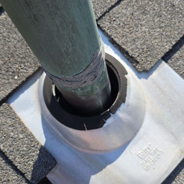 A pipe flashing on a shingled roof, showing a cracked rubber boot seal around the pipe and weathered sealant.