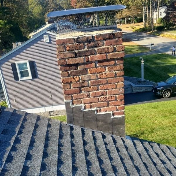 Brick chimney with a metal cap, on a roof with asphalt shingles, partially repaired with black sealant.