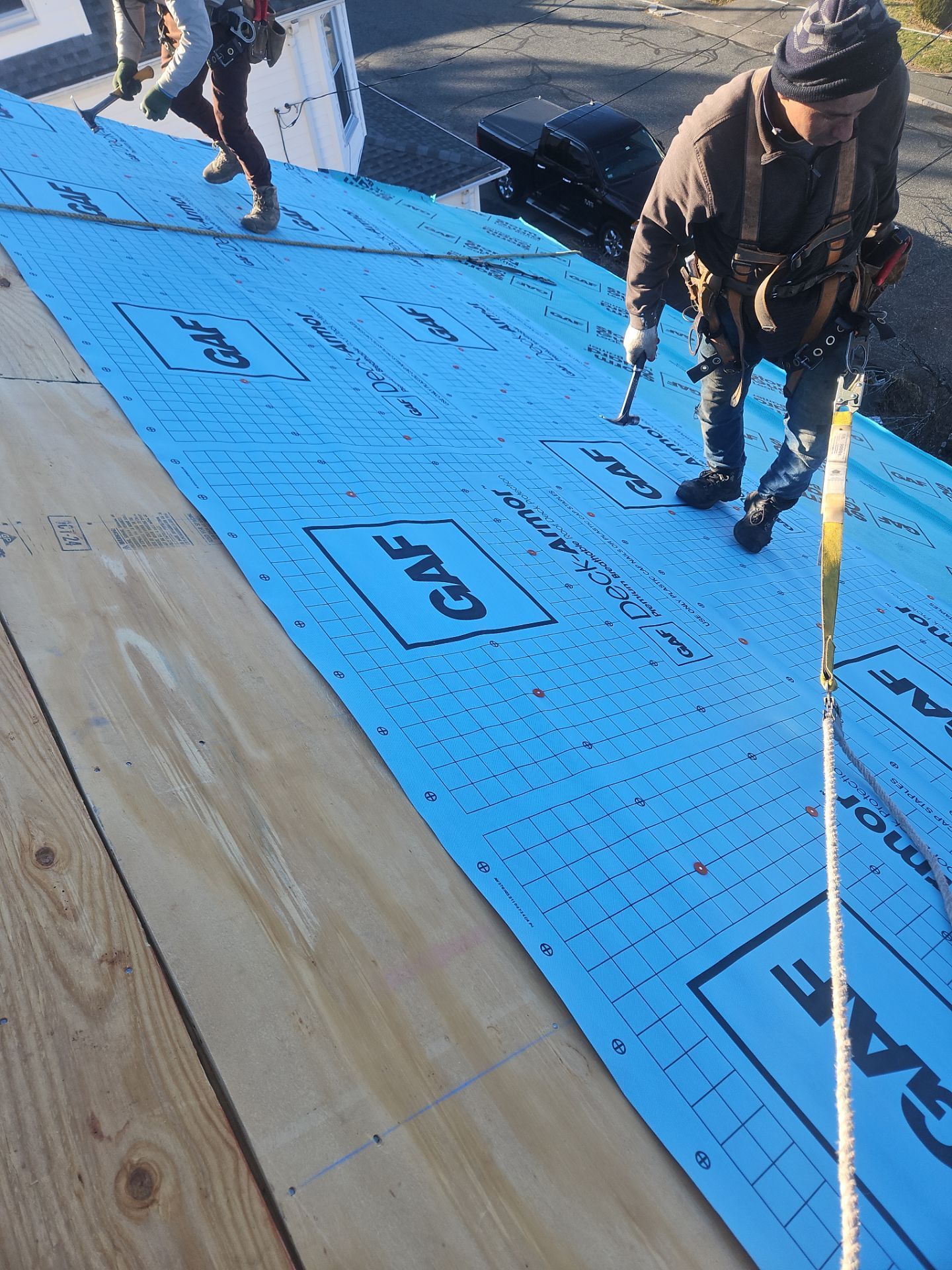 Two workers wearing safety harnesses install blue GAF roof underlayment on a plywood roof deck.