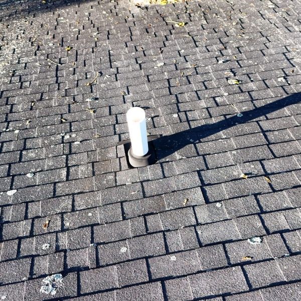 A white plumbing vent pipe emerges through a dark, shingled roof under bright sunlight.