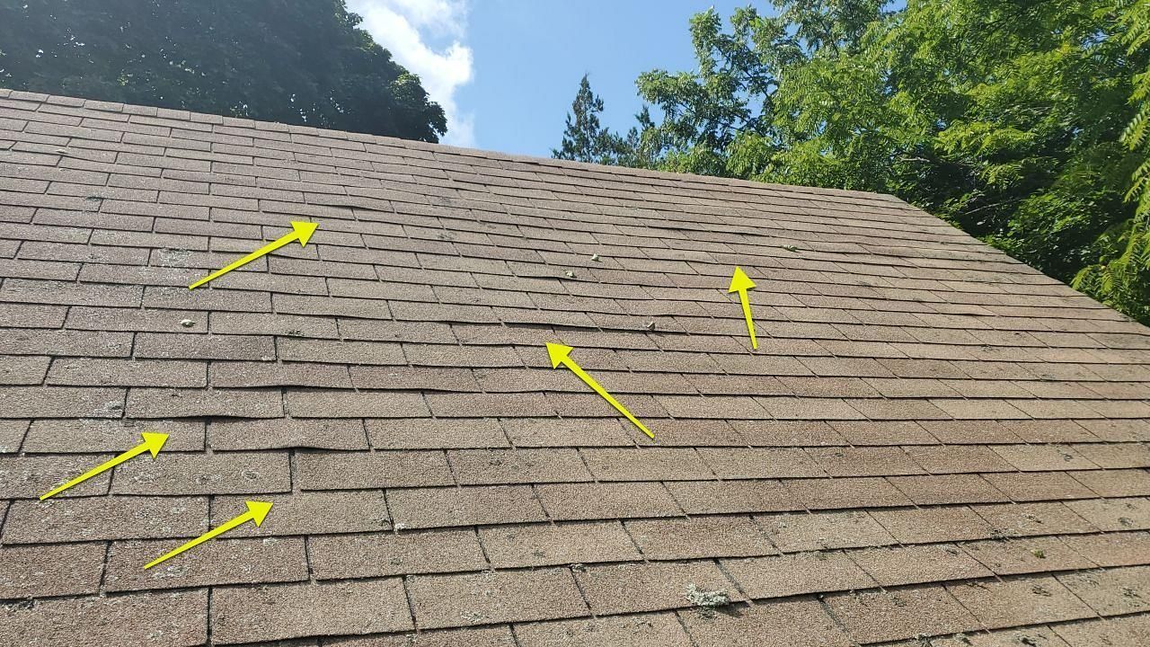 Brown shingled roof with raised shingles, indicated by yellow arrows, under a blue sky.
