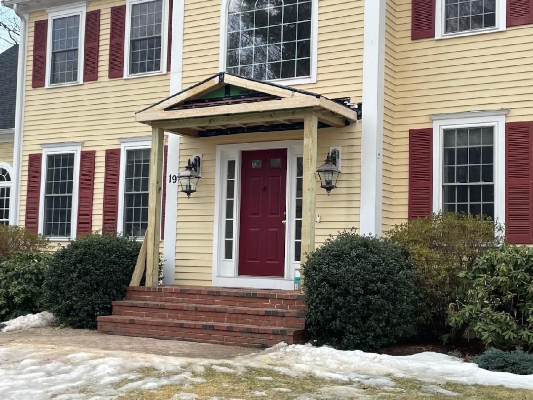 Yellow house with burgundy door, red shutters, and unfinished porch. Snowy ground and bushes.