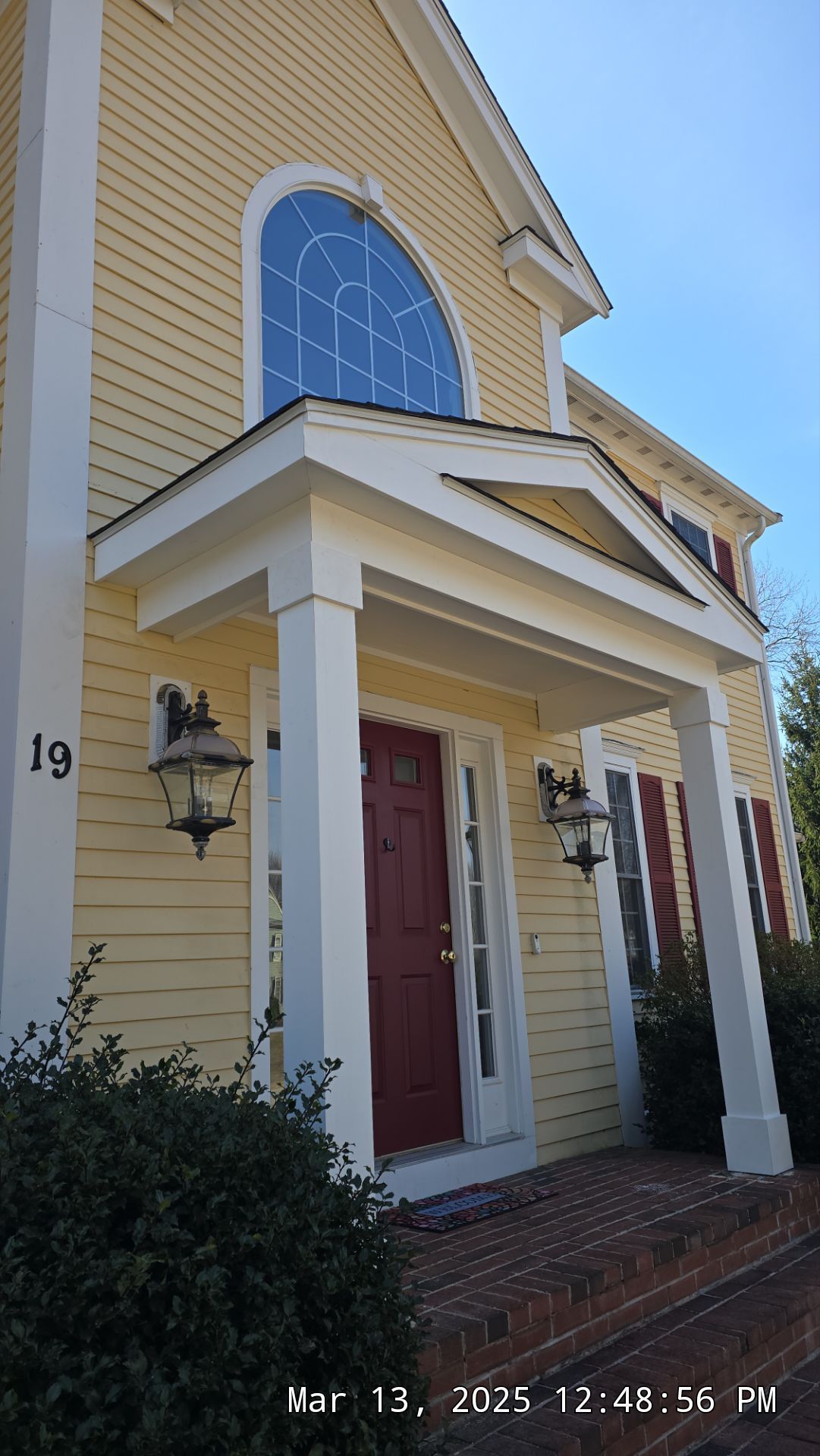 Yellow house with a red door and arched window. White trim and porch. Number 19 on the side.