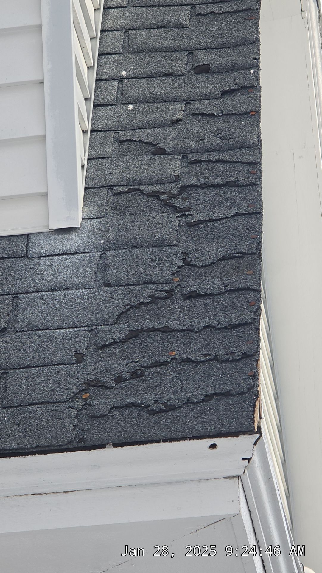 Damaged asphalt shingles on a roof corner, showing curling and missing granules. White trim visible.