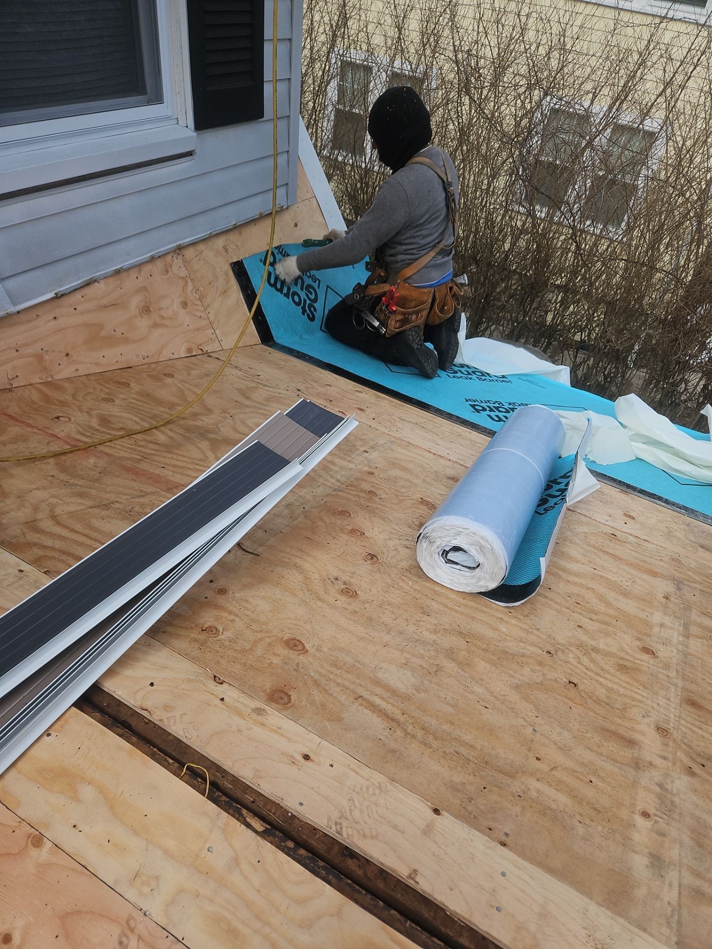 A roofer kneeling on plywood roof, applying blue membrane. Roll of membrane and flashing strips are nearby.