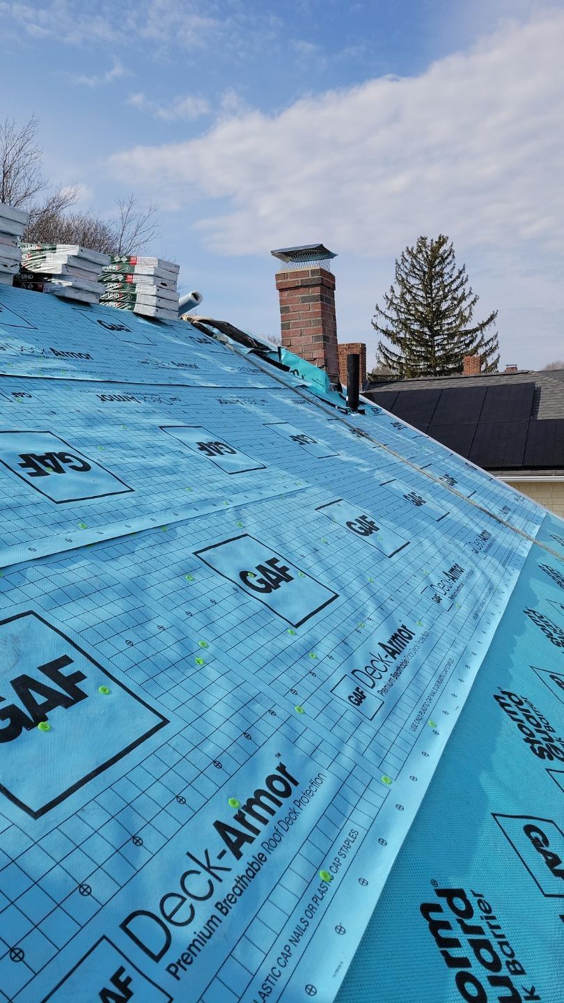 Roof with blue underlayment and stack of shingles, chimney in background.