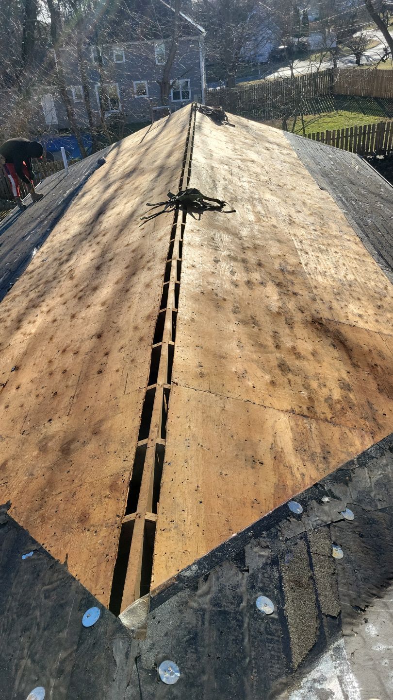 Roofer working on a partially shingled roof. Brown plywood, black shingles, and safety harness visible.
