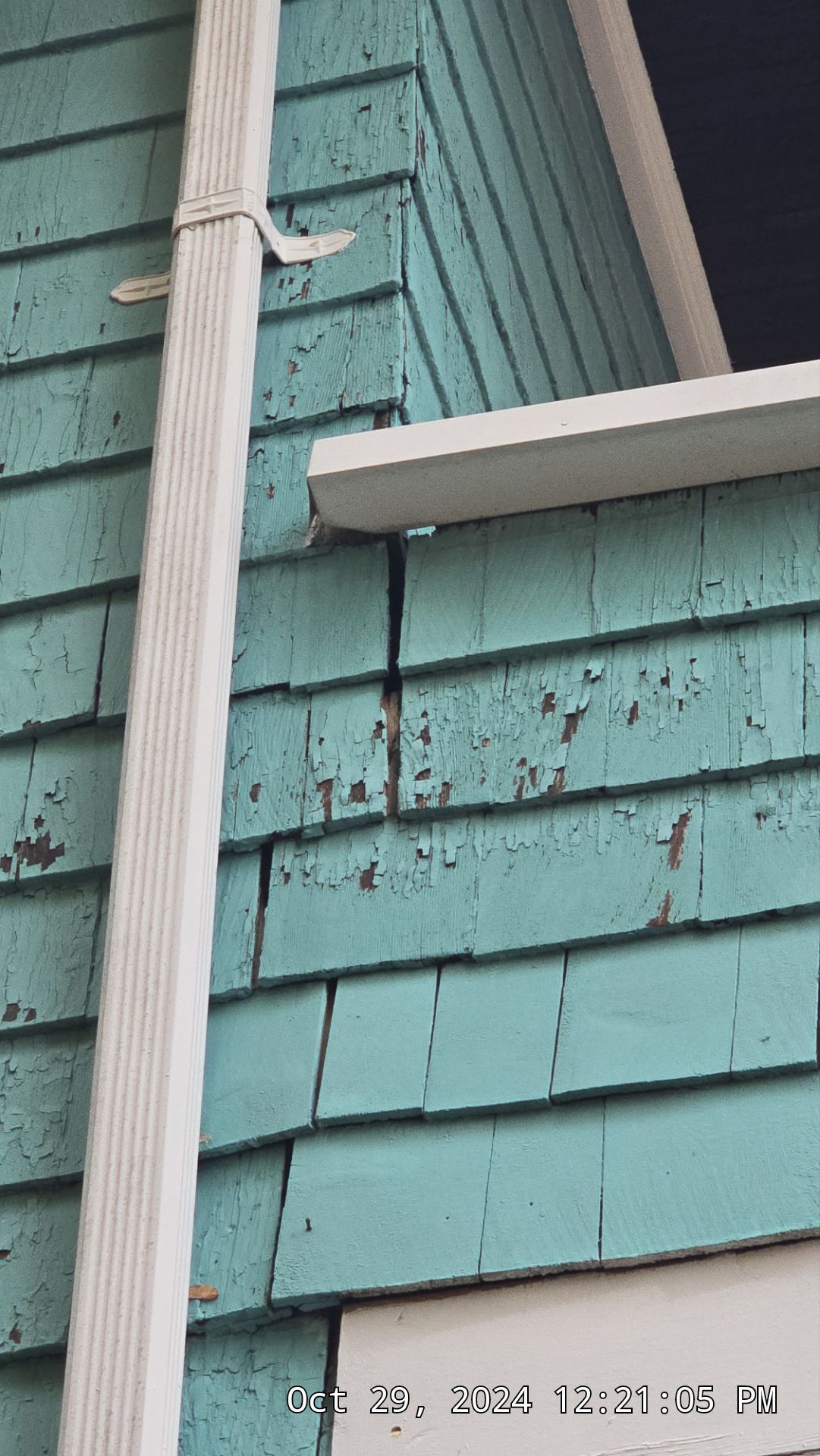 Close-up of a teal-colored house with peeling paint and a white gutter. A black bracket supports a white ledge.