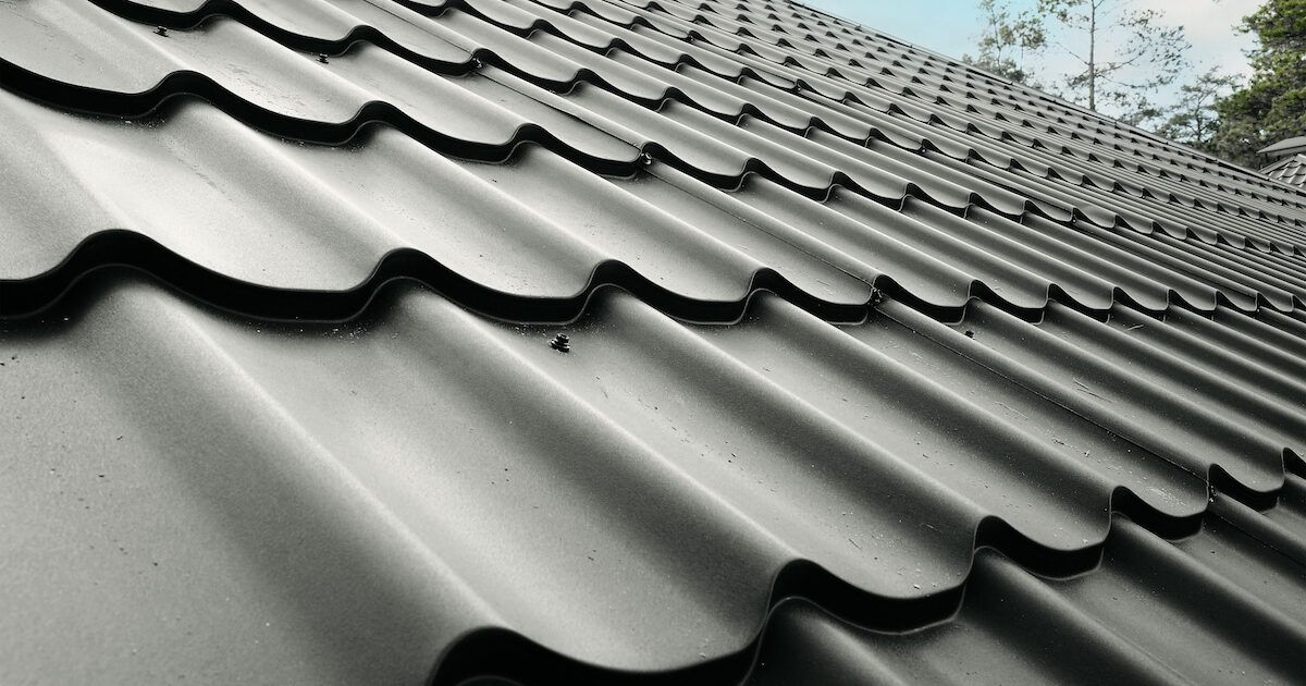 Black metal roof tiles, textured with curved ridges, angled upwards towards a light blue sky.