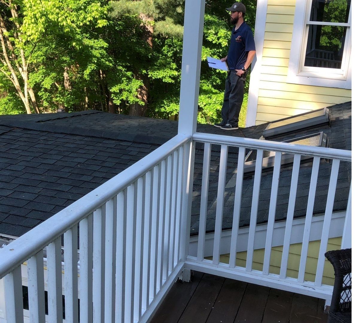 Man on a porch roof inspecting, holding papers. White railing in foreground, trees in background, yellow house.