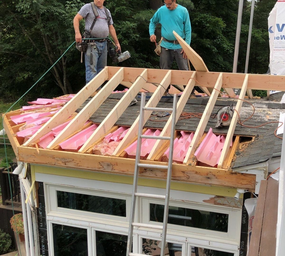 Two workers building a roof, wooden frame with pink insulation. One uses a tool, the other holds wood.