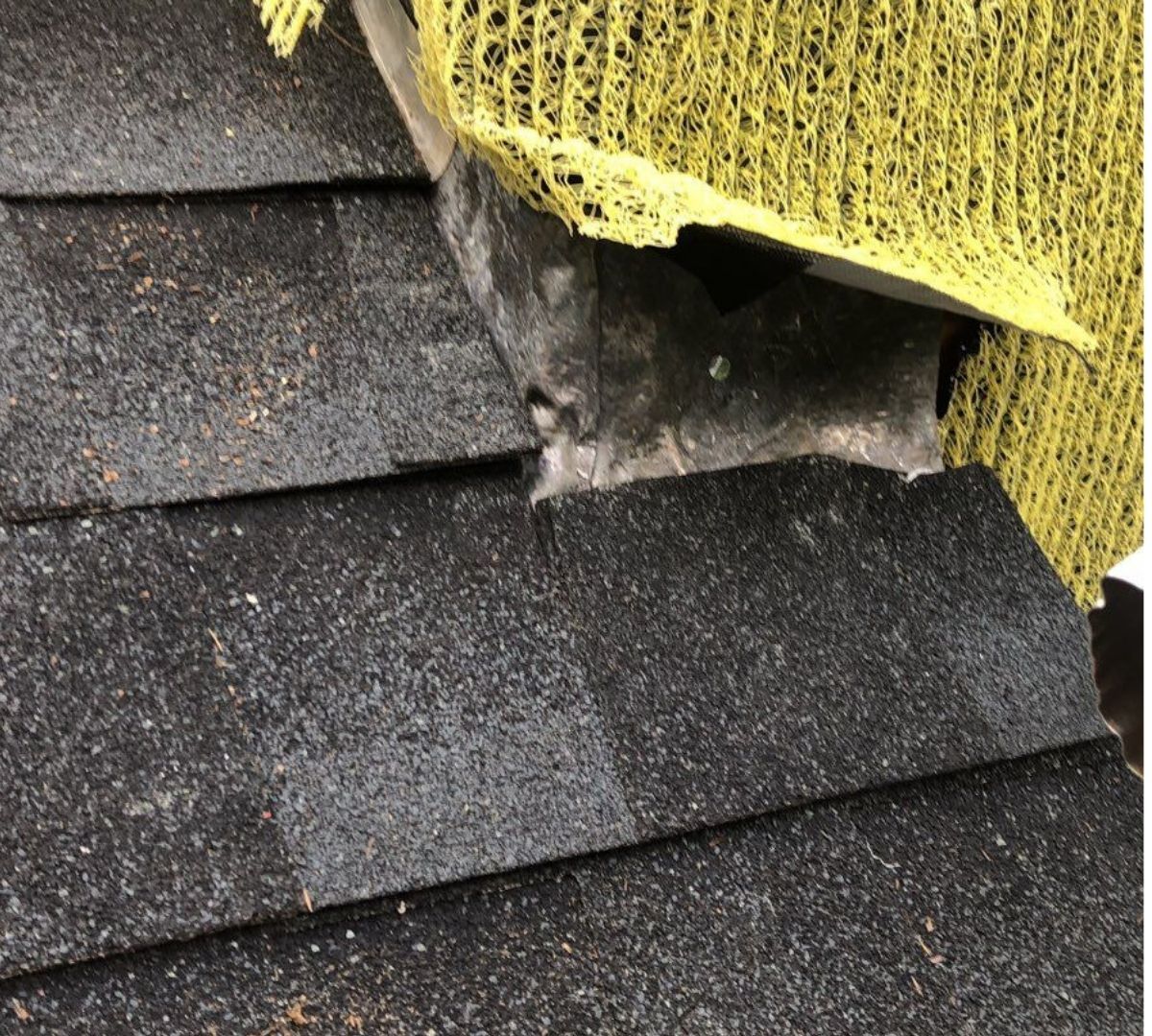 Close-up of a dark asphalt shingle roof with a vent, yellow mesh, and damaged area.
