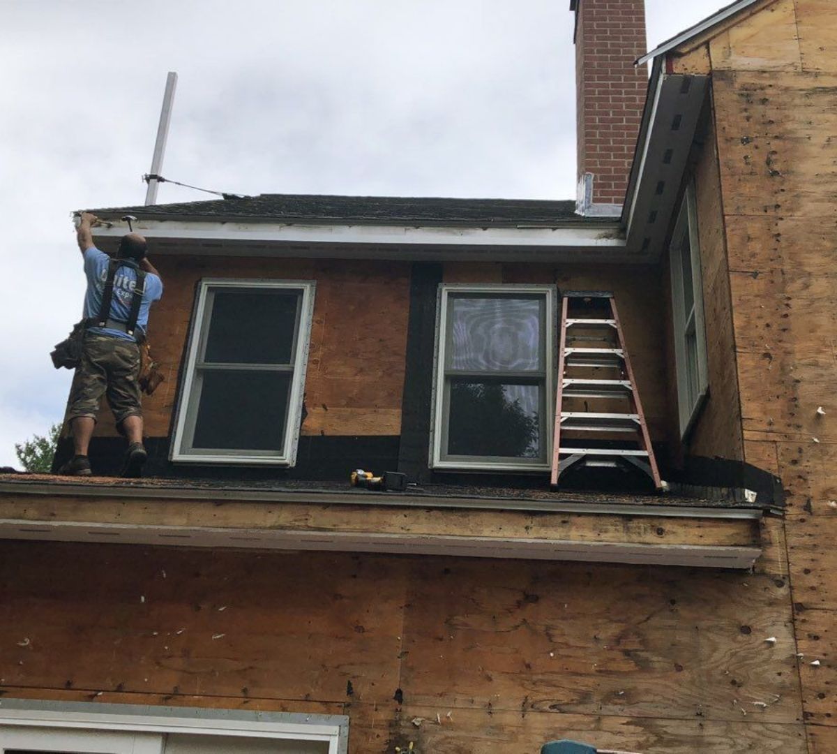 Man on roof repairs siding around windows; ladder present.