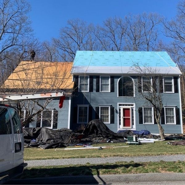 A two-story blue house under renovation with partially replaced roof sections, underlayment, and a worker on the roof.
