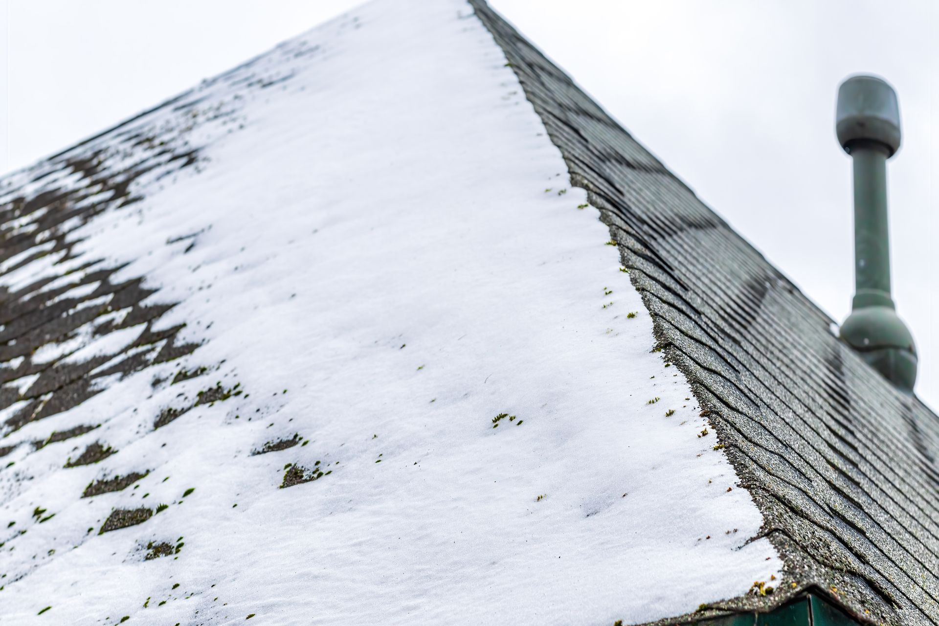 A snow-covered shingled roof with a chimney pipe on the right against an overcast sky.