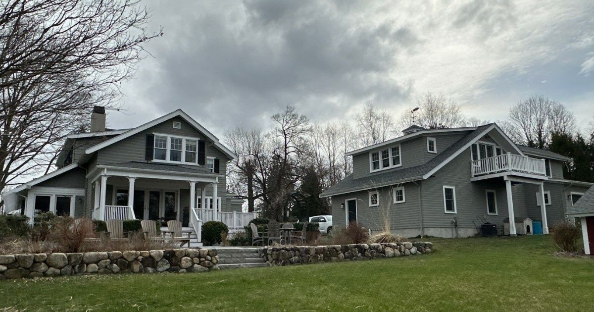 Two gray houses with porches and stone walls on a grassy lawn under a cloudy sky.