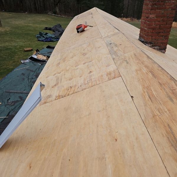 A view from a slanted roof under construction, showing plywood sheathing, a brick chimney, and a nail gun.