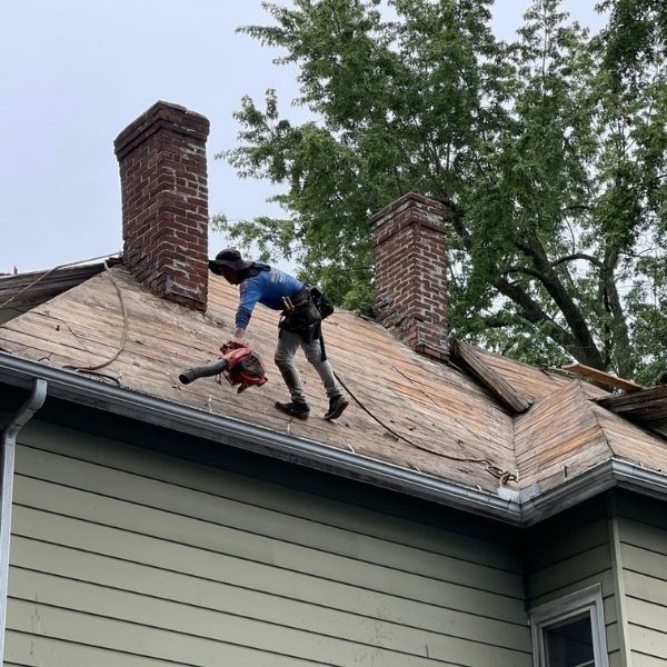 A worker in a blue shirt uses a red leaf blower to clear debris from a wooden roof between two brick chimneys.