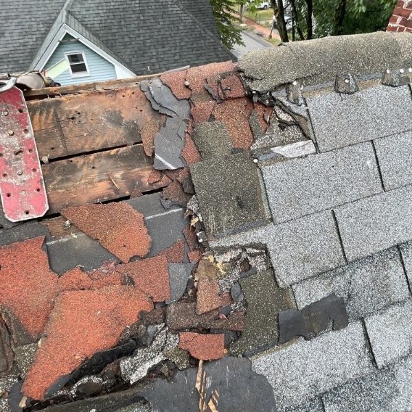 Damaged roof with missing, torn asphalt shingles and exposed wood decking near a metal plate and a brick chimney.