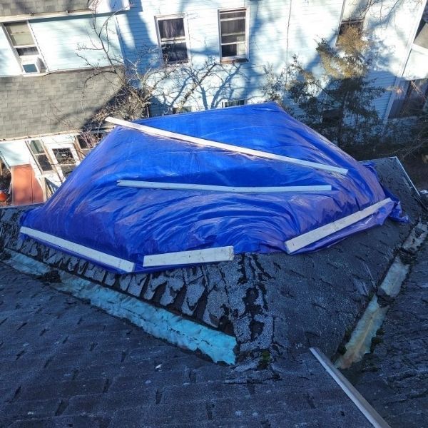A blue tarp covering a damaged section of a residential roof, held down by wooden boards, with shingles in poor condition.