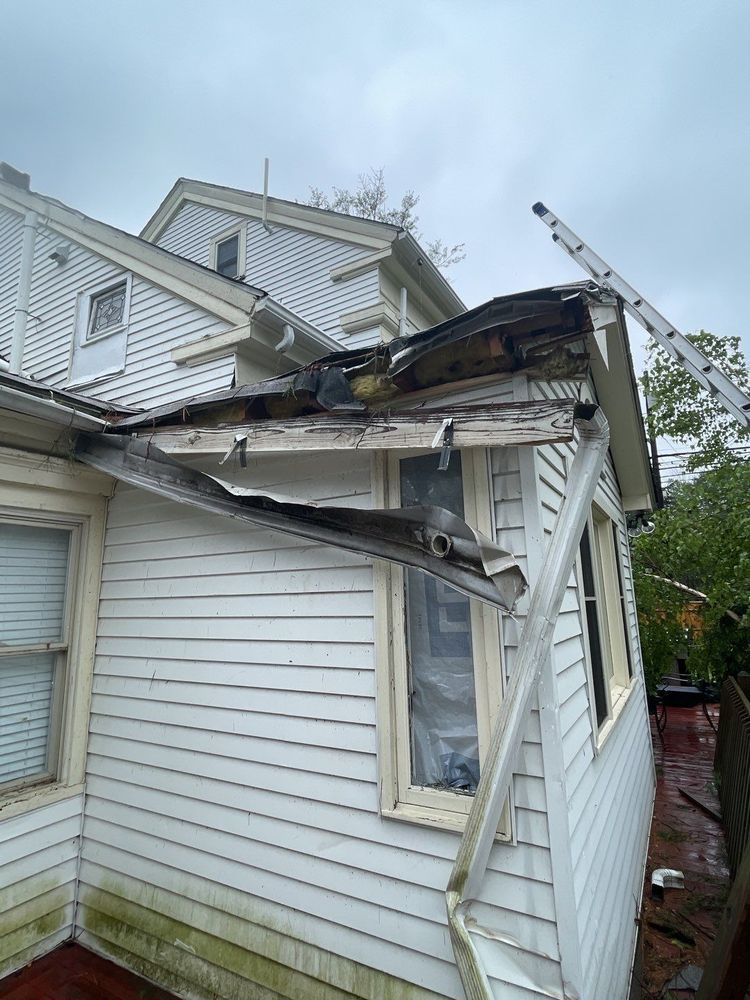 House with damaged roof and siding, likely from a fire. Ladder leans against the roof. Overcast day.