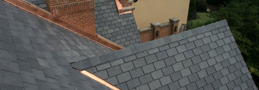 Overhead view of a slate roof with copper gutters and a brick chimney against a building and foliage.