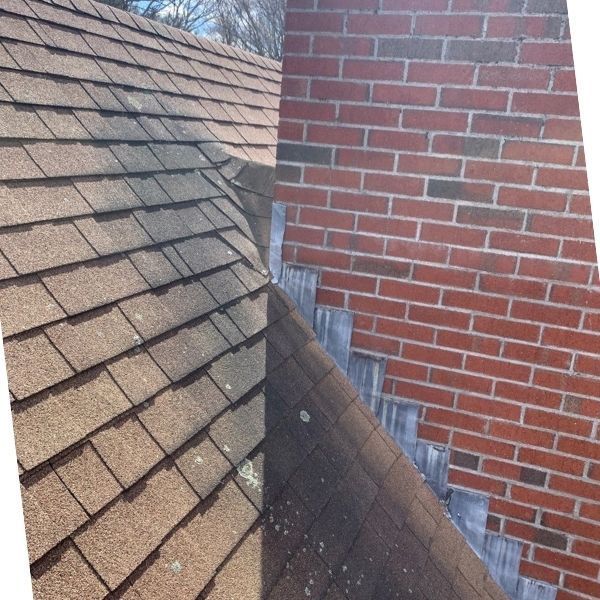 Brown shingles on a sloped roof meet a red brick chimney with metal flashing along the seam.